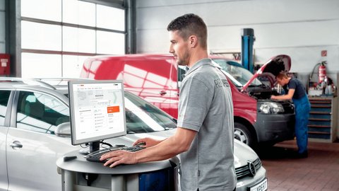 Mann in einer Werkstatt vor einem PC-Bildschirm, neben einem silberenen Auto. Im Hintergrund (unscharf) ein weiterer Mann über einen offenen Motorraum gebeugt. 