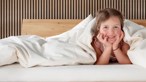 Fotografie eines kleinen Mädchen auf einem Bett, unter einer Bettdecke. Den Kopf auf den Händen aufgestützt und am Lächeln. 