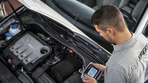 Mann lehnt sich über offenen Motorraum eines Autos. Er hält ein Gerät mit Display in der Hand und trägt ein graues T-Shirt, mit dem Logo REPXPERT" auf dem Rücken" 