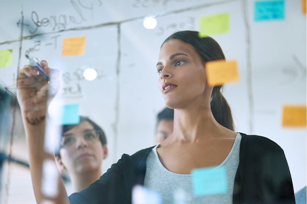 Fotografie von einem Glasboard, mit Post-Its und Notizen drauf. Frau hinter dem Board zu erkennen, die gerade etwas draufschreiben möchte